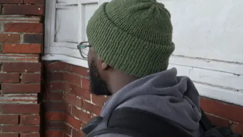 BBC A man wearing a green hat and wearing a grey hoodie and a black rucksack sits facing away from the camera next to a white and brick wall. He has a beard and is wearing glasses.