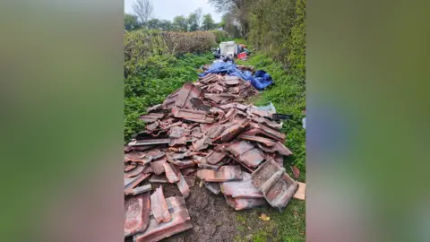 Kitty Fleming Many broken roof tiles are piled on the floor of a grassy bridleway. Further down is a blue tarp covering more waste, and also a white sofa.
