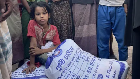 A girl sits on top of a bag containing food aid