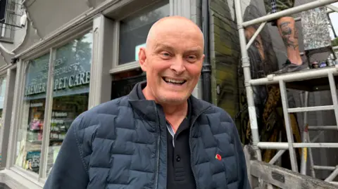 A man with a shaved head is wearing a blue jacket and is smiling. Behind him is a graffiti artist standing on scaffolding as he spray paints a mural on the building behind him. 
