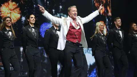 Getty Images Michael Flatley with the cast onstage during the curtain call for the Broadway Opening and debut of 'Lord of the Dance: Dangerous Games' at The Lyric Theatre on November 10, 2015 in New York City. He has blonde hair, wearing a white blazer, red waistcoat white shirt and black trousers. His hands are pointing up to the sides. Dancers are behind him, wearing black tuxedos.