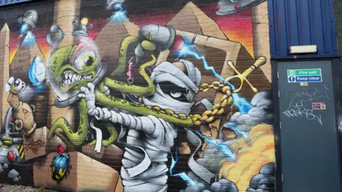 Upfest This picture has a graffiti design next to a fire exit door which reads 'Keep clear'. The design has a cartoon Egyptian mummy fighting an octopus alien with a fish bowl on his head. The mummy is wearing an Egyptian Ank necklace around its neck. The cartoon-styled fight scene has robots and Egyptian pyramids in the background.