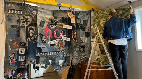 Tom Jackson/BBC A large, handmade rug made up of different pieces of denim jeans hanging from a yellow metal beam in a room. It has various faces on it. Next to it is a woman with her back the camera, who is wearing a denim jacket, white top and black jeans while standing on a ladder trying to attach a different rug - this time a green and patterned one - to the same metal beam and part of the neighbouring wall.