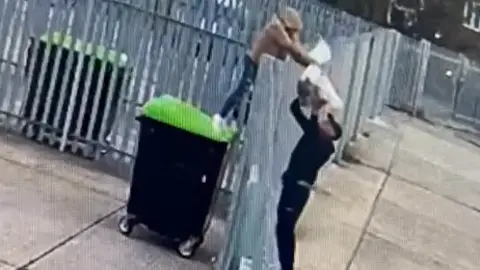 CCTV footage of two figures either side of a metal fence - about two metres high with spikes on toip. One is stood on top of a black bin with a green lid and is passing a clear plastic bag over the fence. The person is dressed in jeans with a brown hoodie. On the other side of the fence is a person with his feet on the ground, dressed in all black with a black cap, and taking the bag above his head.