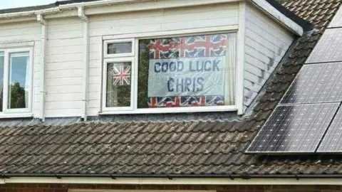 Zoe Millard Upper floor of a house, showing a white-framed window with union jack flags around a banner saying "Good Luck Chris". There are solar panels mounted on the roof to the right.