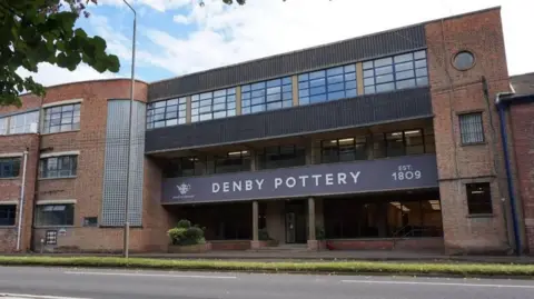 Supplied A large brick building with a hue dark sign which reads "DENBY POTTERY" in white letters.