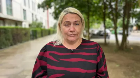 A woman with light blonde hair standing outdoors on a residential street, wearing a black and red striped top, with blurred buildings, trees, and parked cars in the background