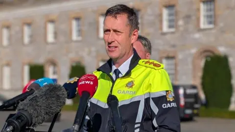 Garda Commissioner Justin Kelly syanding in front of microphones. He has short dark hair, wearing a white shirt, navy tie and high viz garda jacket.