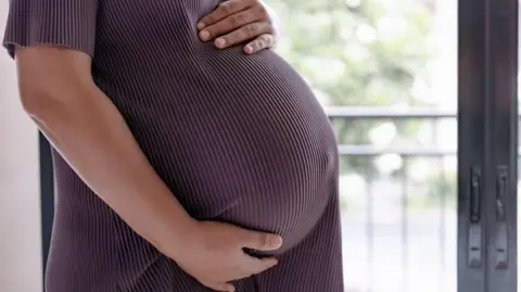 Getty Images A close-up image of a pregnant woman holding her baby bump. Only her bump and hands can be seen and she is wearing a purple dress.