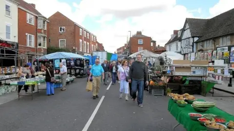 David Dixon/Geograph Archive photograph of Grantham market. There are a number of stalls covered in tarpaulins and people walking along looking at the goods that are for sale.