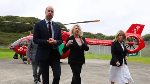 PA Media William walking on the tarmac with two women in front of a red air ambulance helicopter