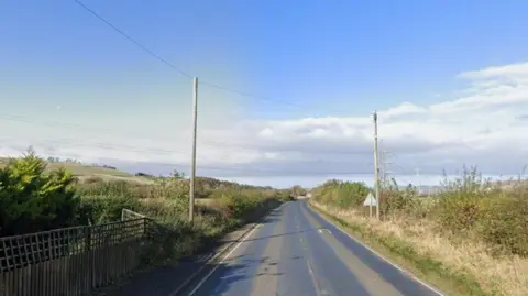A single carriageway country road, with telegraph poles and hedges along the side.