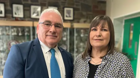 Simon and Barbara pictured in front of school awards cabinet. Simon has short white hair and is wearing thin framed dark glasses. He is wearing a dark blue suit with a white shirt and light blue tie. Barbara has short brown hair with a fringe and is wearing a patterned black and cream shirt with a black top underneath. She is also wearing a gold and silver necklace. 