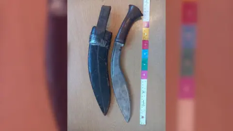 Two long bladed, curved knives on a table top with a tape measure alongside them. 