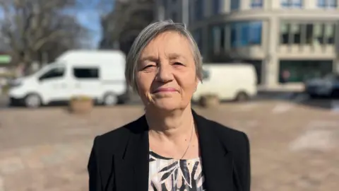 BBC Deputy Mary Le Hegarat is standing in a paved square in front of a road, with an office building behind it. She is wearing a black blazer and is looking at the camera.