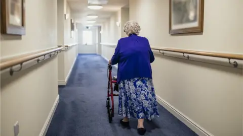 Getty Images A senior woman walking down a corridor with the assistance of a walker