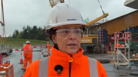 Network Rail Rosario Barcena at the site where the Clifton Bridge is being built. She is wearing a white hard hat, safety goggles and orange high-viz jacket. She is looking in the distance while being interviewed by someone out of shot. She has a microphone pinned to the side of her jacket. Behind her are heavy plant machinery and workmen in high-viz clothing working around the steel structure of the new bridge.