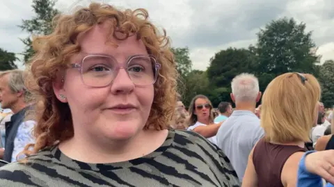 Daisy wearing a black and grey patterned top and large square clear framed glasses. She has ginger curly hair and is looking just off to the right of the camera. In the background there is a large crowd of people and some trees. The sky is overcast and grey.