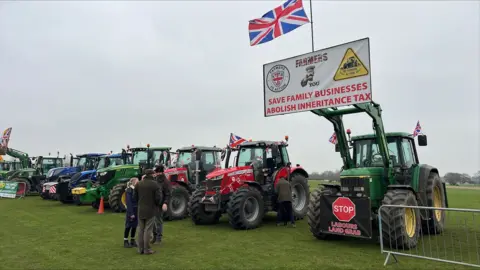 Richard Knights/BBC A green tractor - at the start of a line of them - holds up a white banner saying: Save Businesses, Abolish Inheritance Tax. A group of three people are standing in front of the tractors, parked on grass.