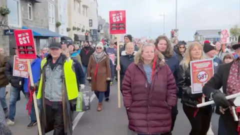A group of people walking along Guernsey's seafront, brandishing anti-GST signs.