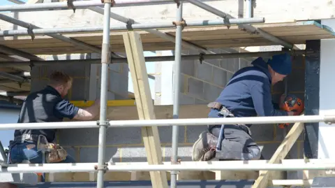 PA Media Two men side-on to the camera are working on a building made of concrete blocks behind scaffolding. One is measuring with a spirit level and one is drilling.