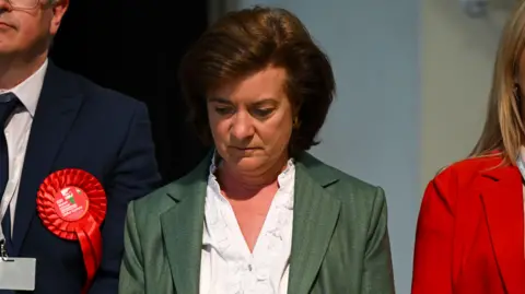 Finnbar Webster/Getty Images Eluned Morgan stood in the middle of two people, looking down at the floor, frowning. She is wearing a green jacket and a white shirt.