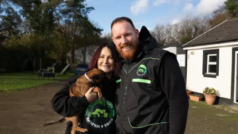 Jacqui and Drren wearing black jacket with their green logo which says Dogs4Wildlife. They are holding one of their Malinois puppies. To the right of them there is a white and black building and to the left of them there are trees and dog training equipment. 