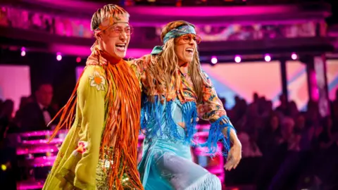 Karen Carney is pictured with a joyful facial expression while dressed in a blue 60s hippy inspired outfit with blue tassles on a pink pattern cropped jacket. Carlos Gu is pictured smiling and is also wearing a green 60s hippy inspired outfit with an orange long tassle scarft around his neck and an orange headband. Both are also wearing round orange framed glasses.