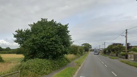 A street view of the B3233. There is a double decker bus in the distance on the right and houses on the right. On the left is a field, bushes and trees.