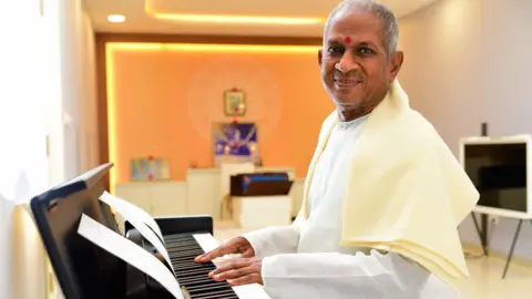 Ilaiyaraaja wearing a white kurta and a pale yellow shawl as he plays a piano