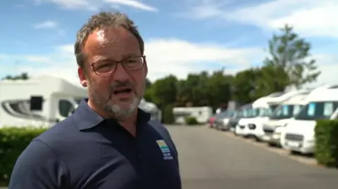 BBC A man with glasses and a beard is wearing blue on the left side of the photo. Vehicles are blurred in the background.