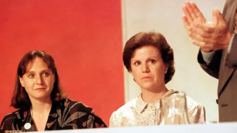 PA Media Rosemary Hunter in patterned brown top and Ann Pearston in a cream top are watching a man in a beige suit on edge of frame talk. They are listening intently. There is a red and white backdrop and some glasses in the extreme foreground.
