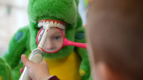 John Fairhall/BBC Child brushing a dinosaur's teeth while holding a magnifying glass over the teeth
