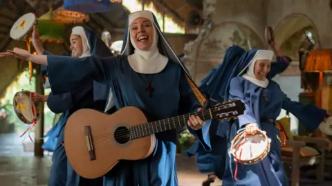 StudioCanal Olivia Colman as the Reverend Mother in Paddington in Peru. She is holding a guitar and has two other women also dressed as nuns next to her.