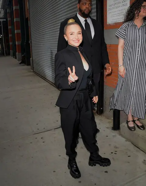 Getty Images Hayden Panettiere, seen on May 19, smiling and posing while doing a peace sign with her fingers on a New York City street. Her blonde hair is tied in a bun and she wears a black blazer, with a black top, black trousers and black boots.