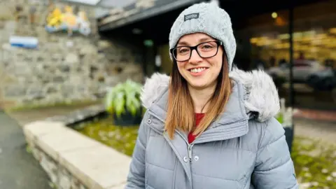 Woman wearing glasses smiling at camera, she is wearing a padded light grey jacket with a furry hood, and a light grey warm hat, standing in from of a library building.