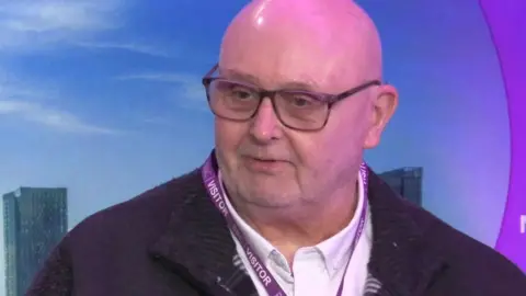 Barry Mackleston, who is bald and has black-rimmed glasses, sits in a purple-coloured BBC Radio Manchester studio as he speak to a presenter. 