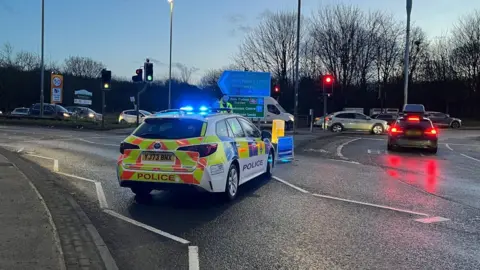 Police car with blue lights flashing stands at entrance to Stanningley Bypass in Leeds, with a queue of traffic in the background.