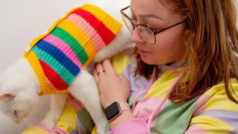 Charly Allso A woman in a striped hoodie with a white cat walking from her shoulder down her arm, also wearing a rainbow striped pullover. The woman has red hair and is wearing glasses and a nose stud.