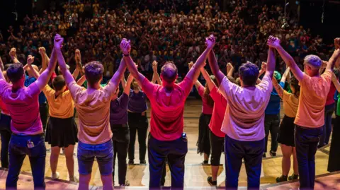 Soul Media Dozens of people in colourful T-shirts stand on a stage at the Bristol Beacon, linking hands as they salute the audience who are applauding in the background