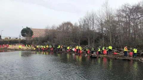 BBC About a hundred figures in high-vis yellow vests. They are litterpicking and carrying red bags. They are along the edge of the River Taff. 