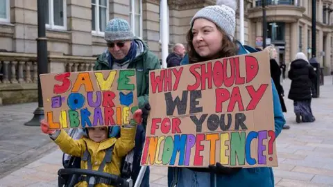 Getty Images A man, woman and child in a pram stand with colourful home-made signs at a protest against council cuts. The signs read "Save our Libraries" and "Why should we pay for your incompetence?"