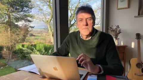 A man sitting at a wooden desk with his laptop open and a woodland scene in the background.