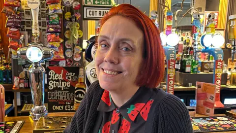 A woman with short red hair, wearing a black top with strawberries on it is sat in front of a bar 