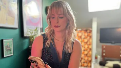 Jess sitting in her flat looking at her phone. A TV on a cabinet can be seen behind her, with a turquoise wall with framed artwork on her left.