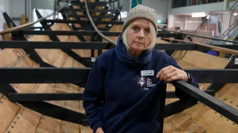 Joe Barett/BBC Jacq Barnard stands within the reconstructed ship's hull. One arm rests on black metal framework as she smiles at the camera. She is wearing a grey beanie hat, blue neck snood and a navy jumper with a name badge on her chest. 