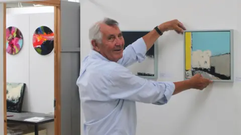 Lansdown Art Studios Association Mike, smiling to camera, holding up a small abstract painting in turquoise, grey and yellow