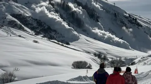 Three people watch in foreground as avalanche barrels down mountainside ahead, from left to right