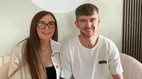 Two people sitting on a cream sofa. The woman, on the left, has long hair and black-rimmed glasses. The man has short hair cut in a fringe and a white T-shirt.
