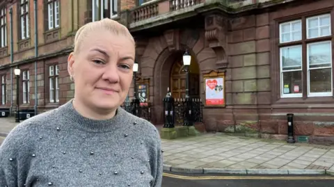 Andrew Turner/BBC Emma Flaxman-Taylor has long blonde hair, tied back in a pony tail. She is wearing a grey jersey which has silver bobbles sewn on. She is standing outside Great Yarmouth Town Hall, a red-stone building dating to 1883 which features decorative masonry, sash windows and a large oak double door.